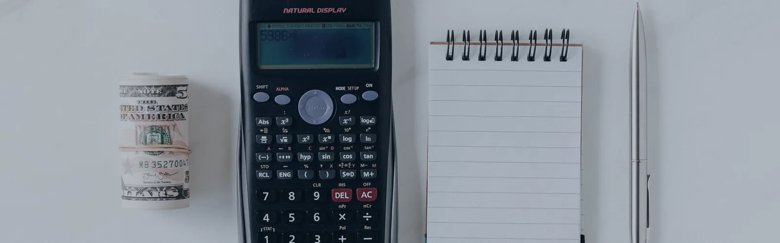 Calculator, rolled banknotes and a blank notepad on a white desk